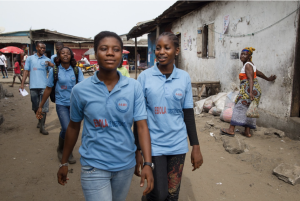 A group of young people in blue shirts walking in a village area with rustic buildings.